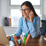 Single happy female business owner with smile and eyeglasses on phone and working on laptop computer at desk with bright window in background